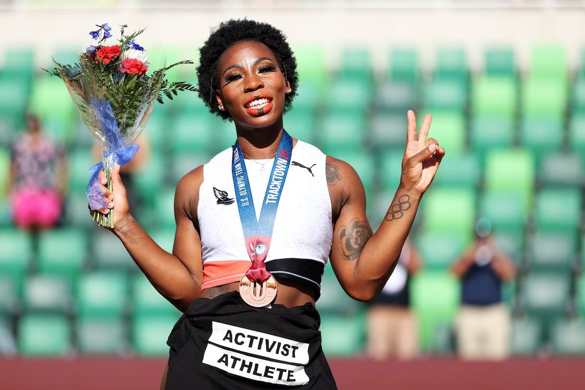 Gwen Berry smiles holding a bouquet of flowers and flashes a peace sign, wearing a bronze medal around her neck and a shirt tied around her waist reading "activist athlete"