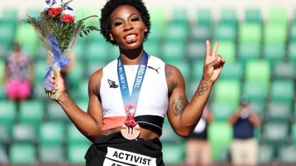 Gwen Berry smiles holding a bouquet of flowers and flashes a peace sign, wearing a bronze medal around her neck and a shirt tied around her waist reading 