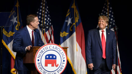 Donald Trump stands on a stage wearing wrinkled pants, smiling at NC Senate candidate Ted Budd