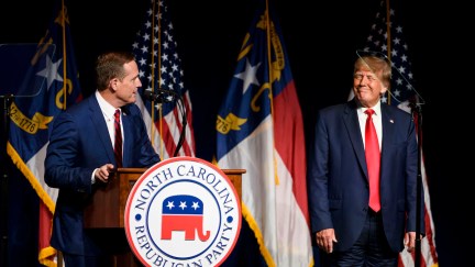 Donald Trump stands on a stage wearing wrinkled pants, smiling at NC Senate candidate Ted Budd