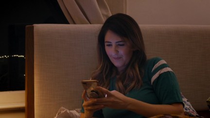 D'Arcy Carden sitting in bed, looking at her phone in Ride the Eagle.
