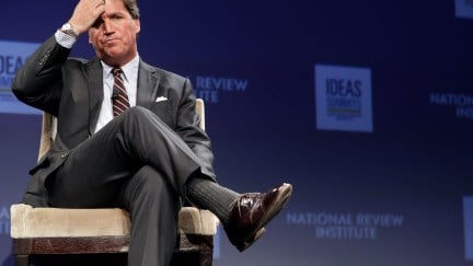 Tucker Carlson sits cross-legged, scratching his head during a talk at a convention