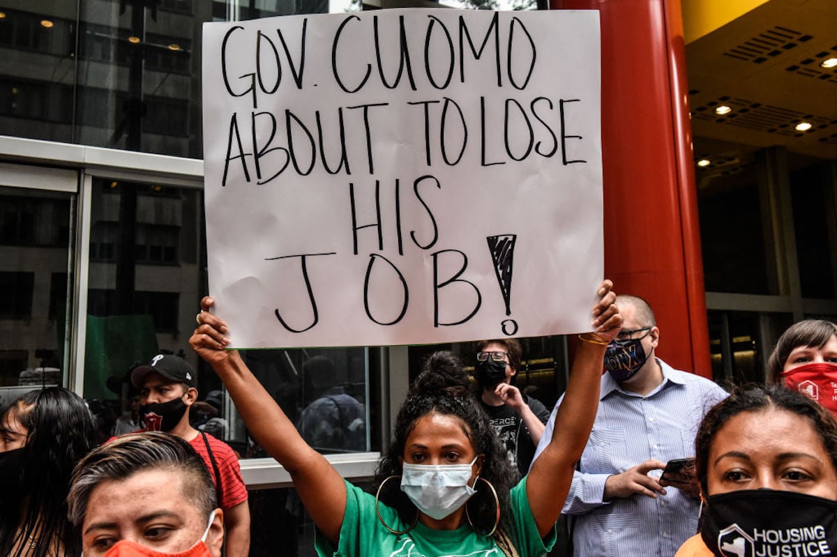 A protester wearing a mask holds a sign reading "Gov. Cuomo about to lost his job!"