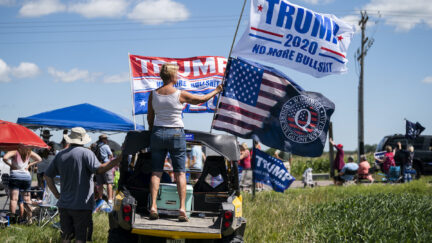 A small crowd gathers outside with a woman stands on the bed of a pickup truck holding a Trump 2020 flag and a Q Anon flag