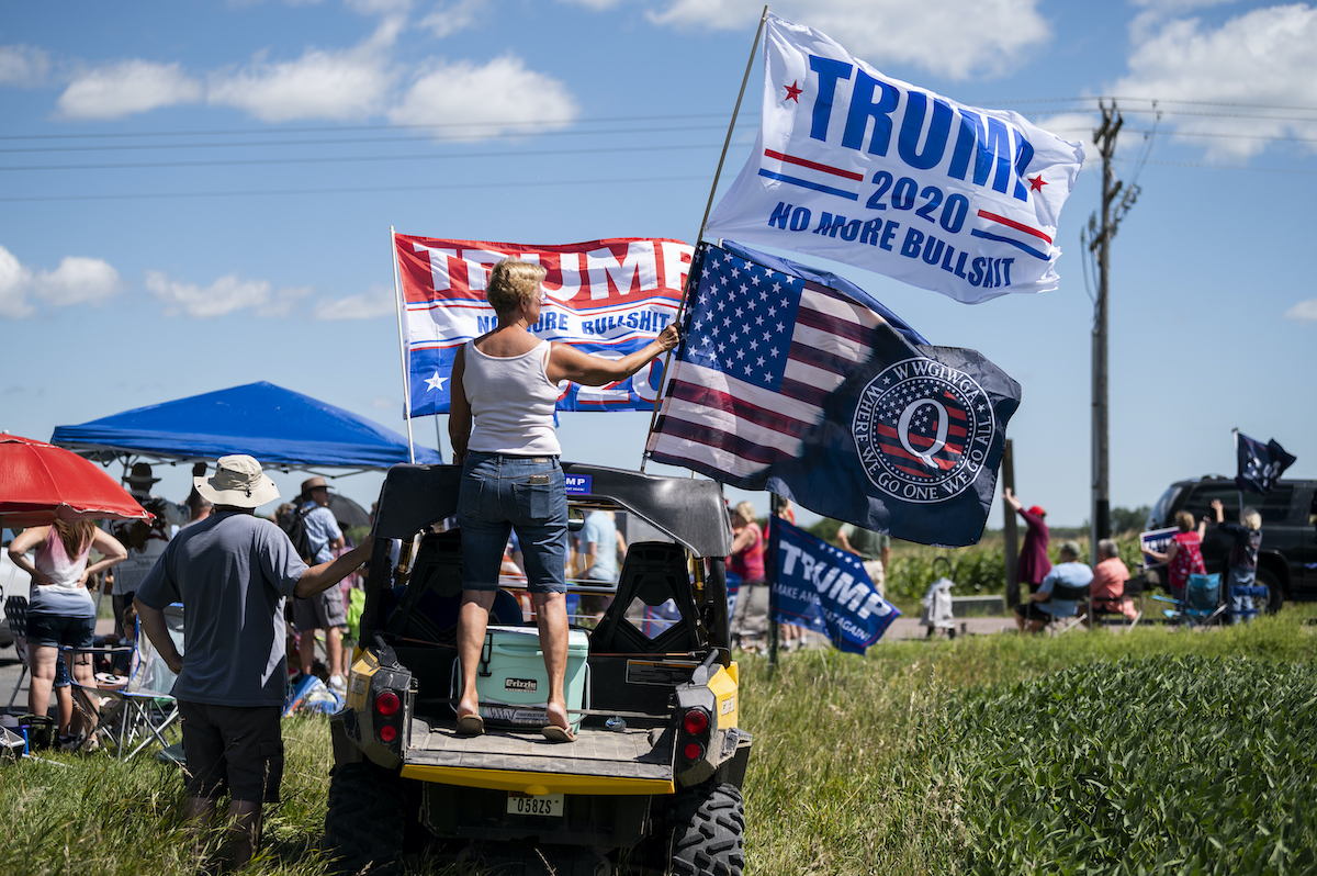 A small crowd gathers outside with a woman stands on the bed of a pickup truck holding a Trump 2020 flag and a Q Anon flag