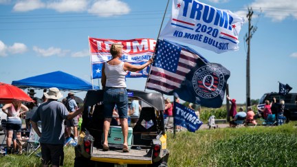 A small crowd gathers outside with a woman stands on the bed of a pickup truck holding a Trump 2020 flag and a Q Anon flag