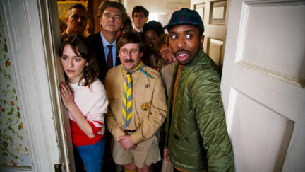 A crowd of people looks into a room through a doorway in a scene from Ghosts