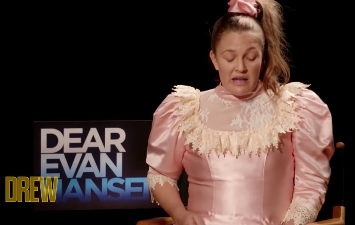 Drew Barrymore in character as Josie Geller from Never Been Kissed, wearing an 80s pink satin prom dress , against a black background with the words Dear Evan Hansen behind her.