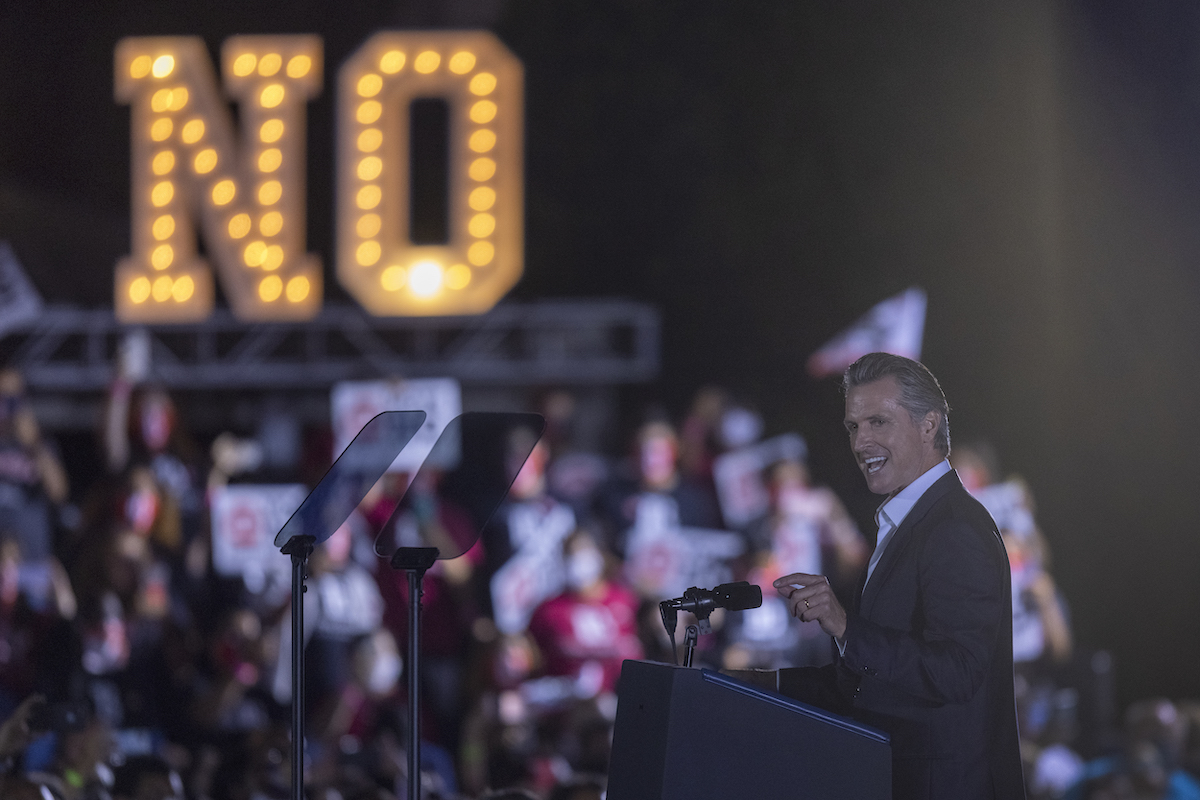 At an election rally, Gavin Newsom speaks to a crowd in front of a giant sign made of lightbulbs spelling out "NO"