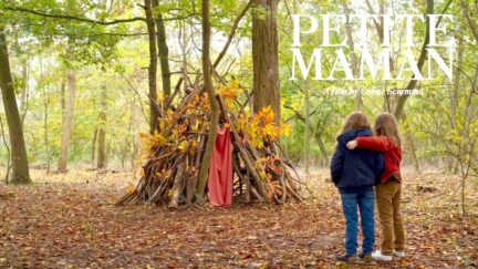 Two girls embrace as they observe a shelter they've built in the woods in Celine Sciamma's Petite Maman.