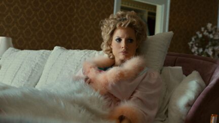 Jessica Chastain wears a pink feathered robe and full makeup lying in bed, looking angry as Tammy Faye Bakker in 'The Eyes of Tammy Faye'