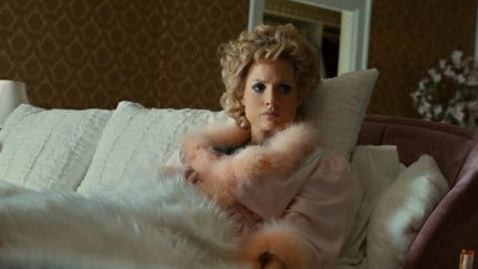 Jessica Chastain wears a pink feathered robe and full makeup lying in bed, looking angry as Tammy Faye Bakker in 'The Eyes of Tammy Faye'