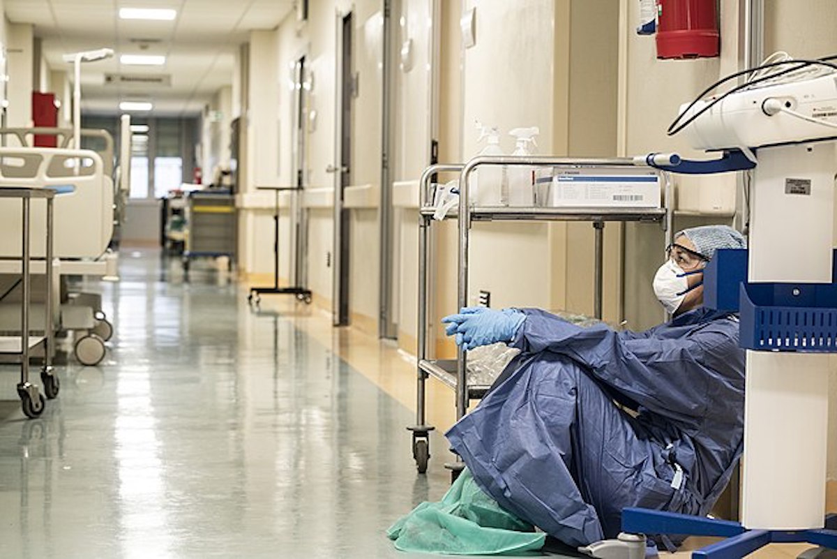 An exhausted healthcare worker sits wearing a mask and gown on a hospital floor