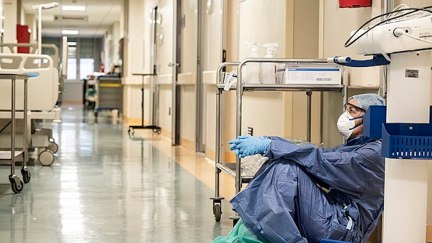 An exhausted healthcare worker sits wearing a mask and gown on a hospital floor