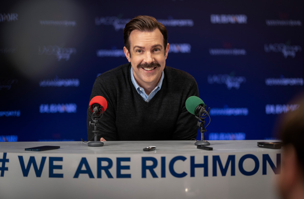 Ted Lasso smiles during a press conference in an episode of Ted Lasso