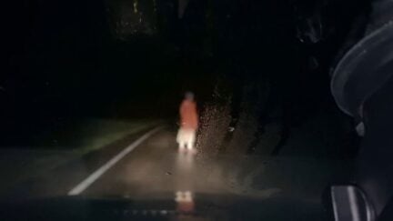 A blurry person in a red coat is seen on a dark road through a windshield in horror movie 'Dashcam'