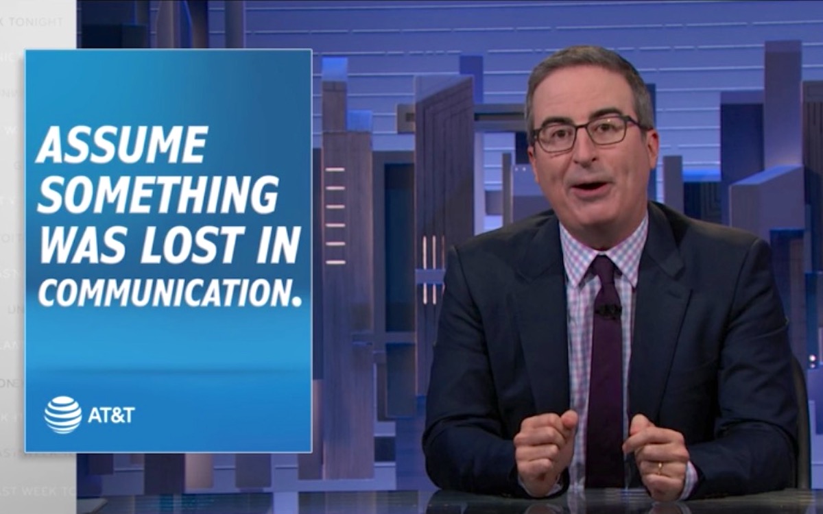 John Oliver speaks from his newsdesk during an episode of Last Week Tonight next to a graphic reading "Assume something was lost in communication - AT&T"