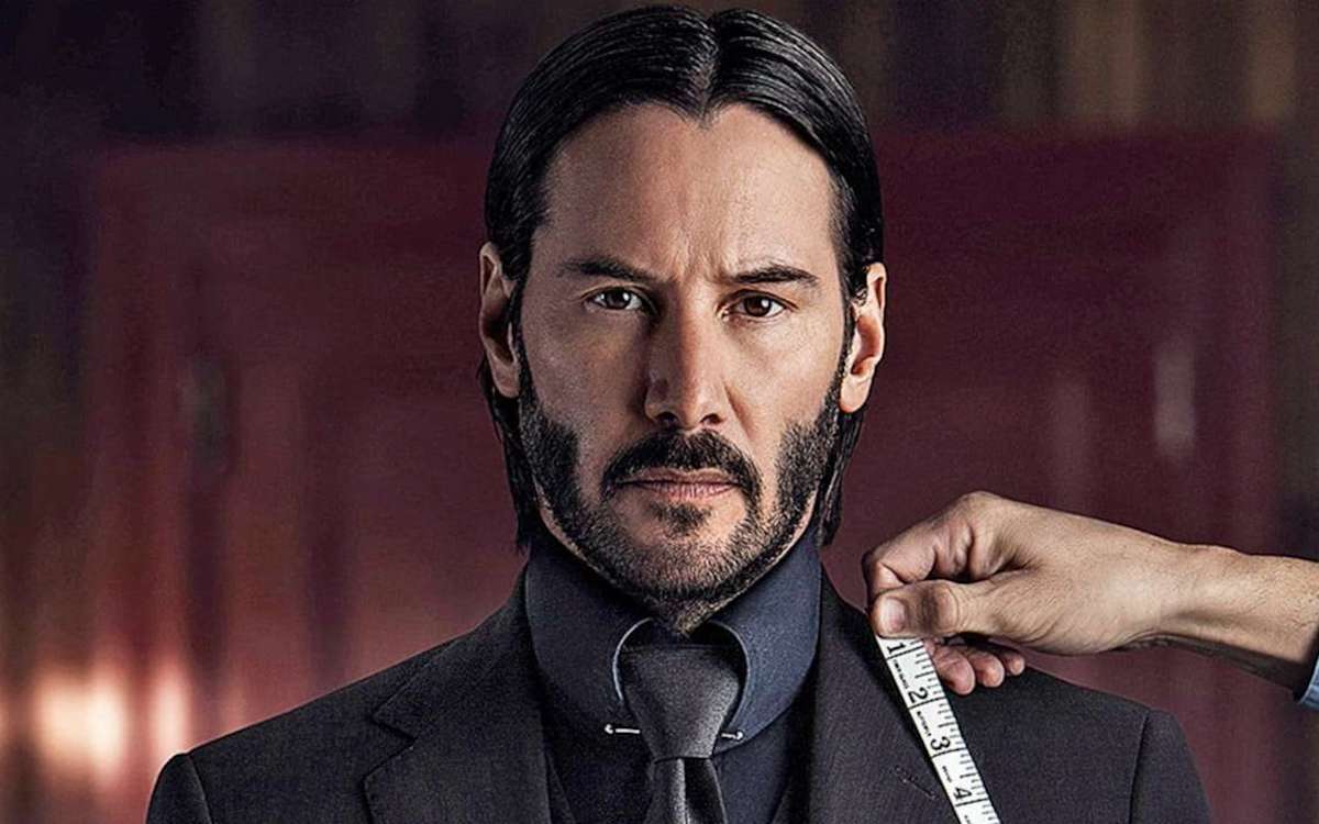 Keanu Reeves as John Wick looks into the camera as a tailor's hand holds a measuring tape to his collar