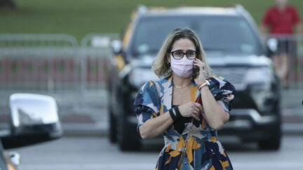 Kyrsten SInema wears a mask and a bright floral dress, talking on a cell phone in a parking lot