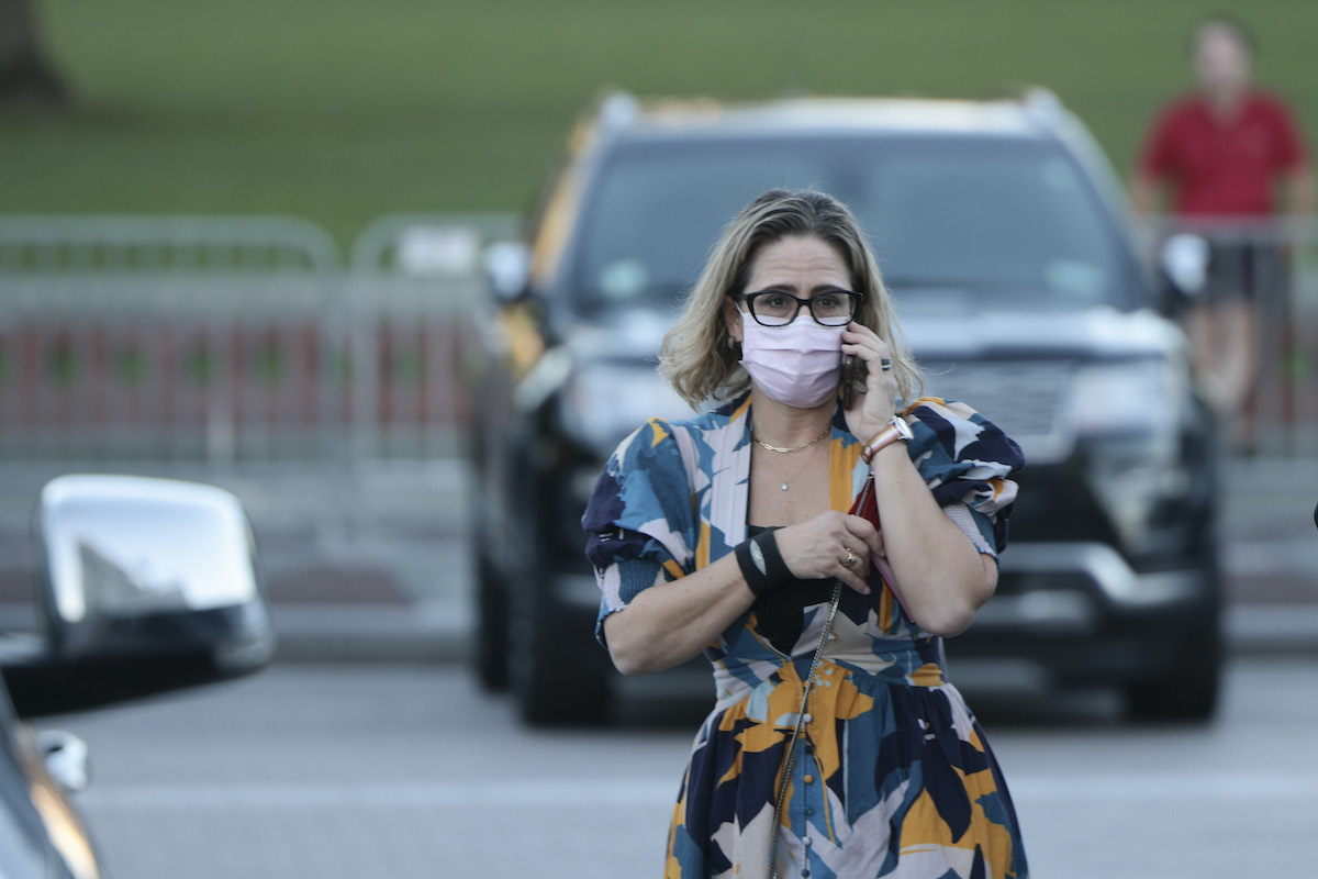 Kyrsten SInema wears a mask and a bright floral dress, talking on a cell phone in a parking lot