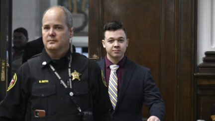 Kyle Rittenhouse looks into the camera and smirks while walking into the courtroom behind a guard.