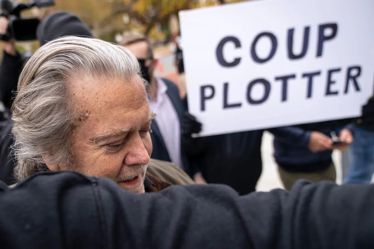 Steve Bannon arrives at an FBI office. Someone in the crowd behind him holds a sign reading "COUP PLOTTER"
