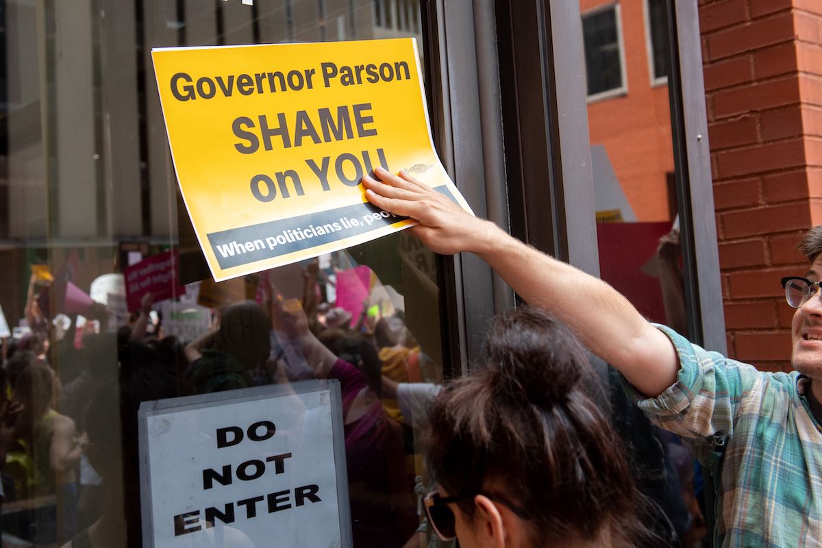 A protestor holds up a sign to a glass door reading "Governor Parson shame on you"