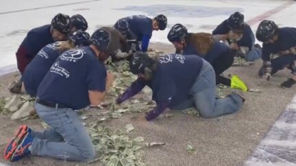 Ten people in matching blue t-shirts crawl on hands and knees, grabbing at a pile of dollar bills.