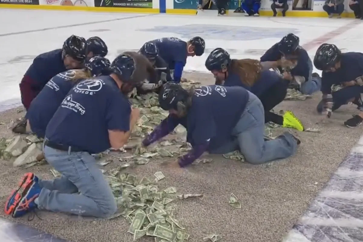 Ten people in matching blue t-shirts crawl on hands and knees, grabbing at a pile of dollar bills.