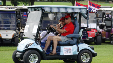 Two older people ride on a golf cart decorated with Trump flags.
