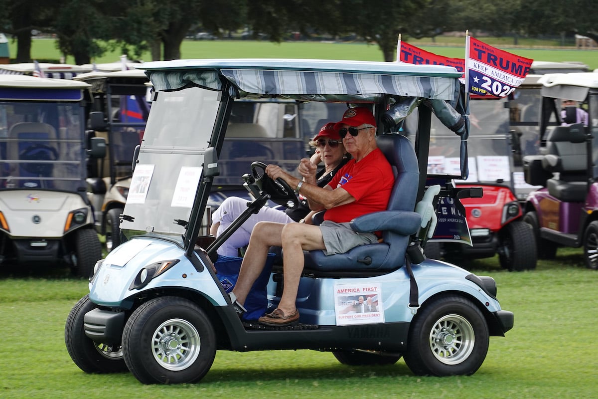Two older people ride on a golf cart decorated with Trump flags.