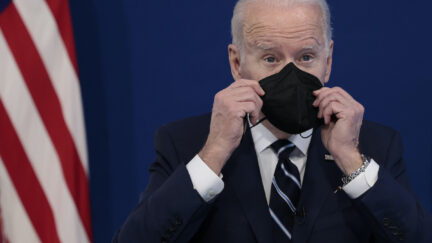 Joe Biden holds a black face mask up to his face during a press conference in front of an American flag.
