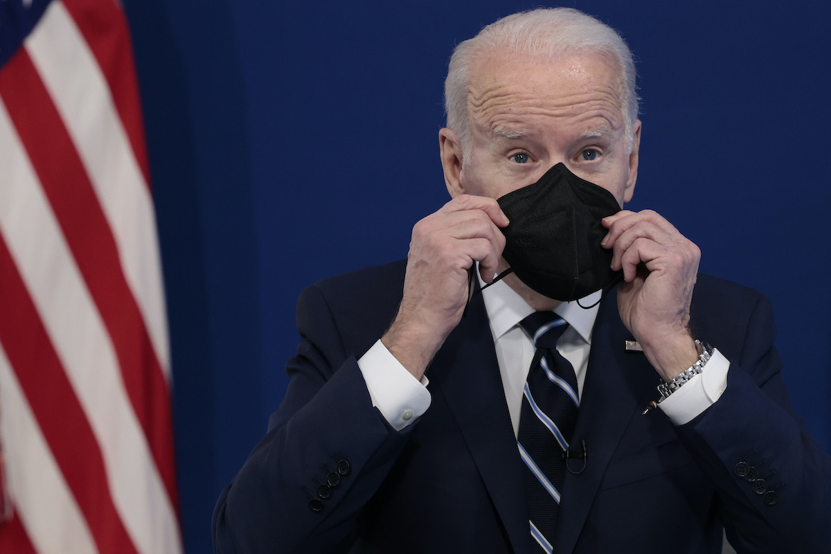 Joe Biden holds a black face mask up to his face during a press conference in front of an American flag.