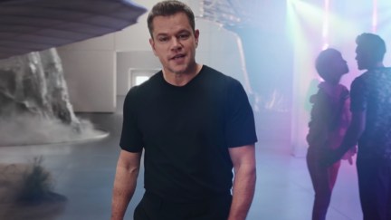 Matt Damon walks on a film set past an old timey airplane on his right and a couple flirting on his left.