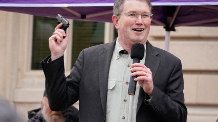 Thomas Massie holds up a small gun and smiles while holding a microphone during a rally.