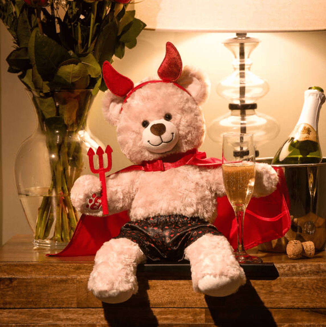 A teddy bear wearing devil horns, a cape, and satin boxers, sitting next to a glass of real champagne.