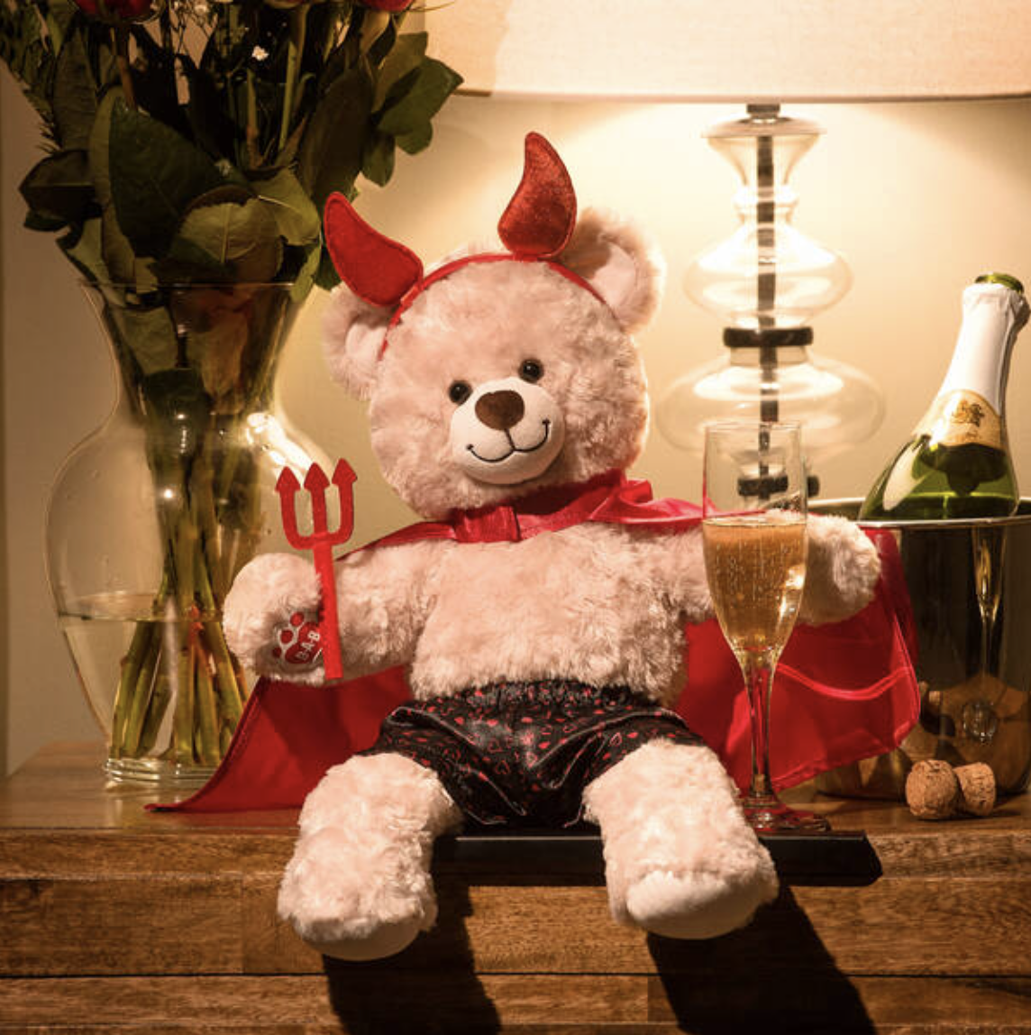 A teddy bear wearing devil horns, a cape, and satin boxers, sitting next to a glass of real champagne.