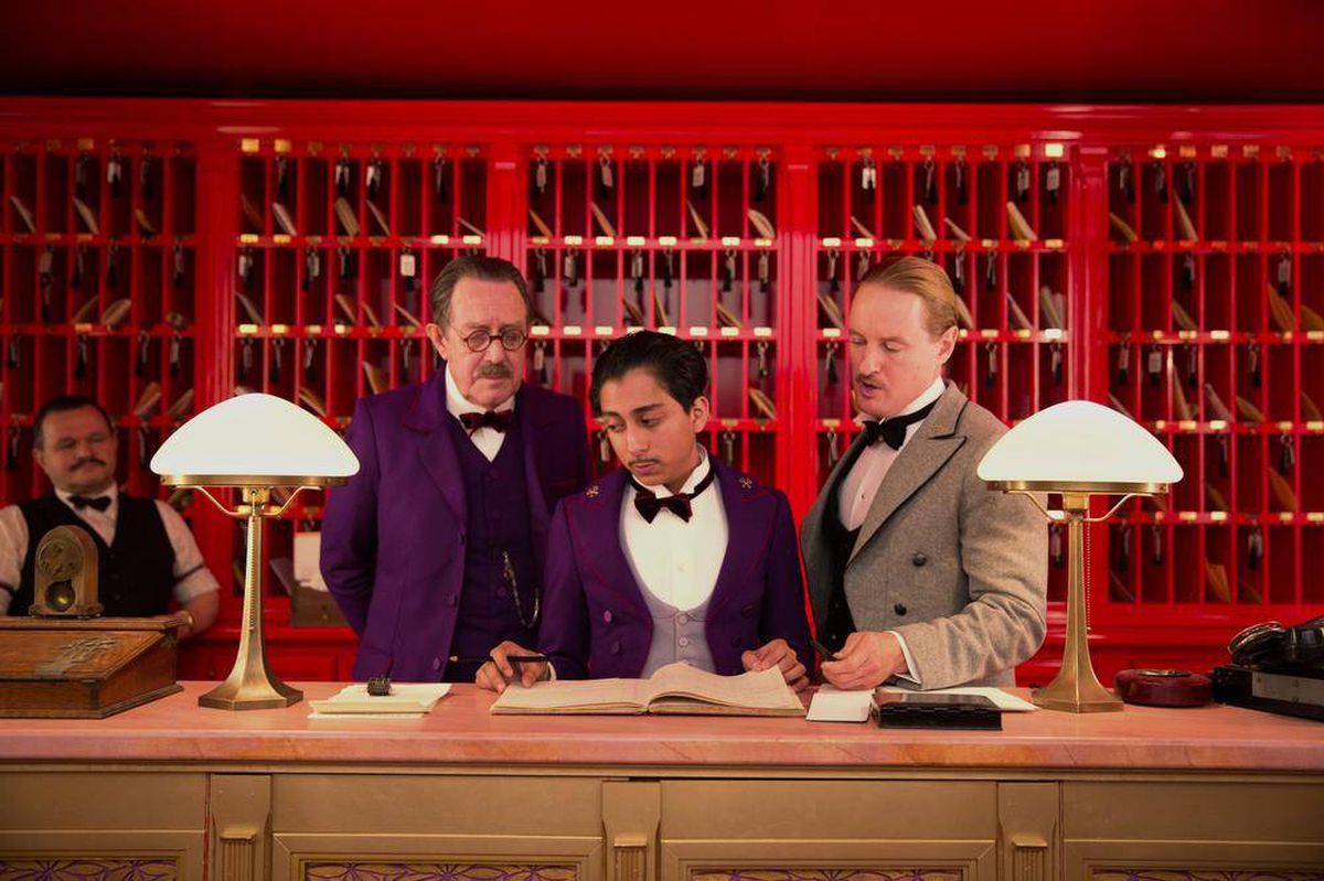 Three men including a bellboy stand at the front desk of a hotel, in front of an all red background with mailboxes and room keys in "the Grand Budapest Hotel."