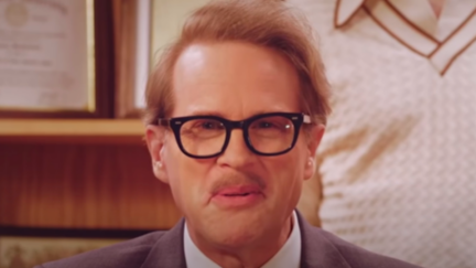 In a scene from The Hyperions, Cary Elwes wears thick glasses and speaks into the camera.