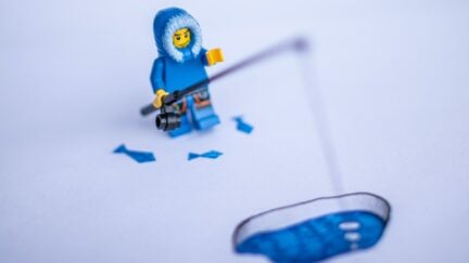 A lego minifig wearing a parka holds a fishing pole into a hole in ice