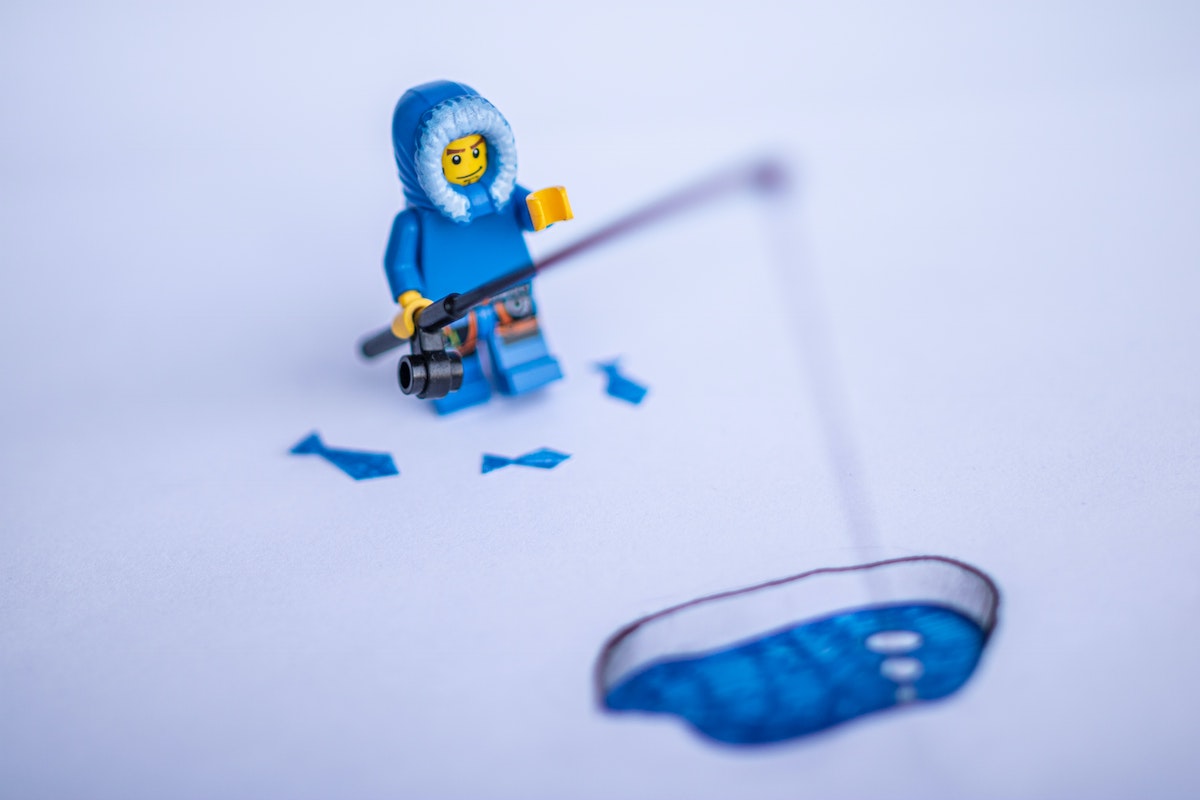 A lego minifig wearing a parka holds a fishing pole into a hole in ice