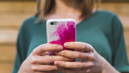 A white woman with her face obscured holds a cell phone in a pink case out in front of her