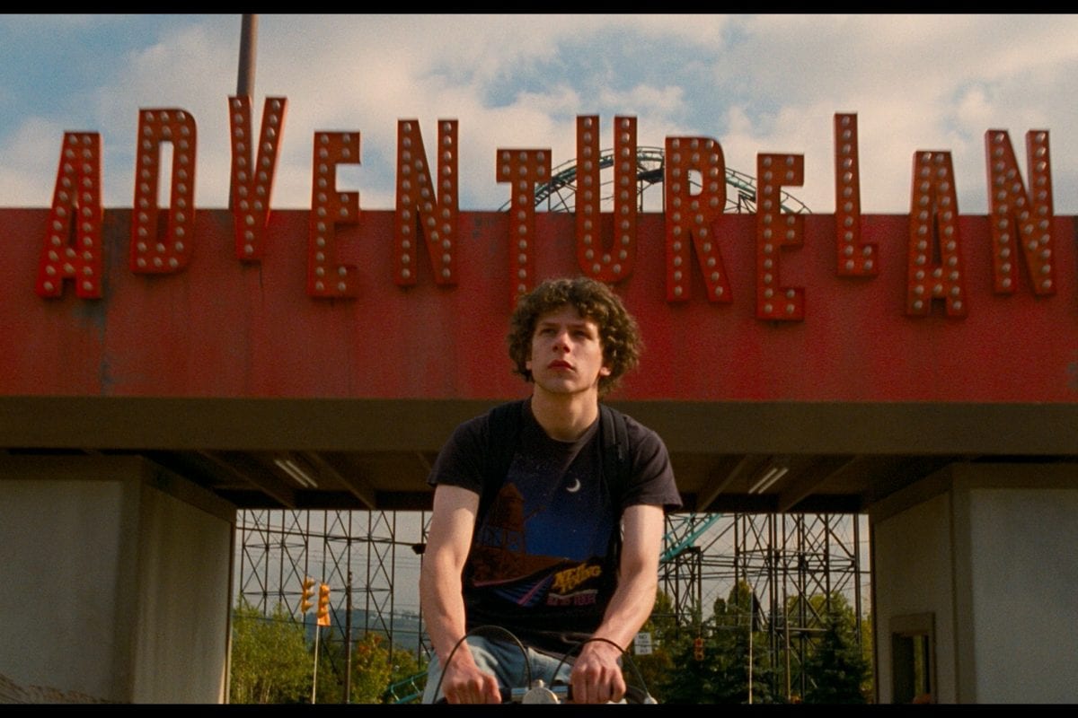 Jesse Eisenberg in front of theme park, Adventureland, sign