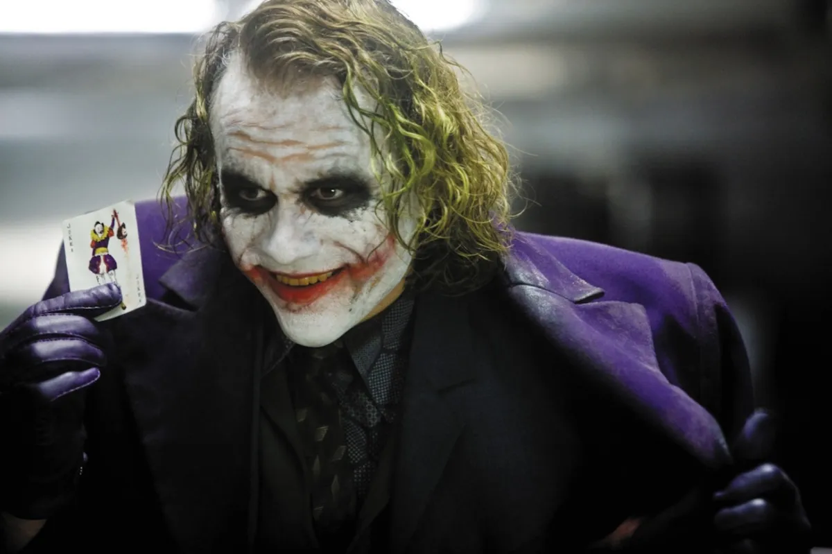 Heath Ledger as the Joker holding up a card in Batman: The Dark Knight.