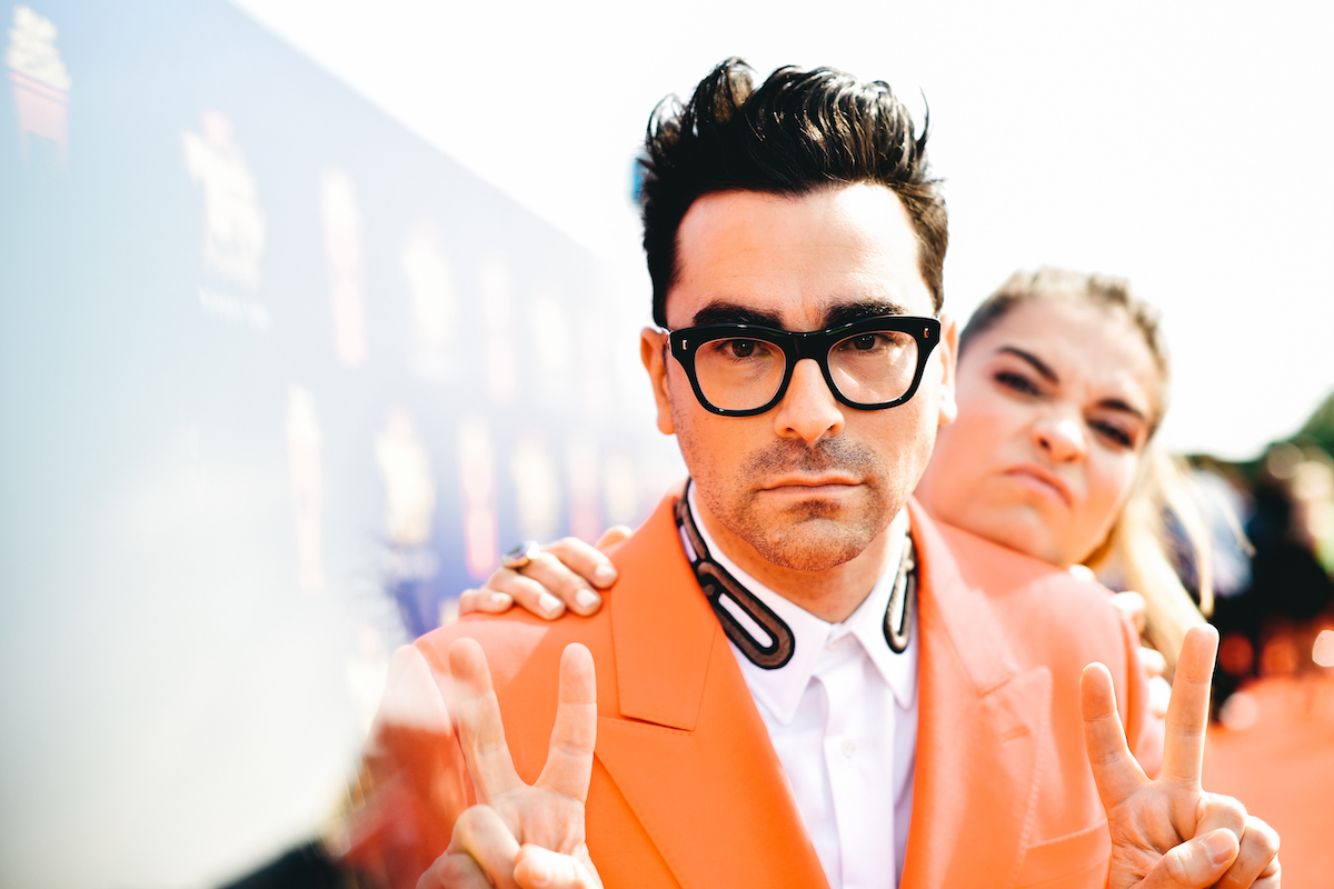 Dan Levy wears an orange suit, looks into the camera and flashes two peace signs while Annie Murphy leans over his shoulder and scrunches her face