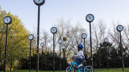 A young child rides a bike through a park featuring an art installation of many clocks on tall poles.