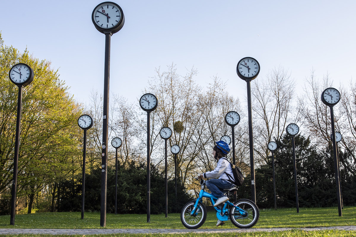 A young child rides a bike through a park featuring an art installation of many clocks on tall poles.