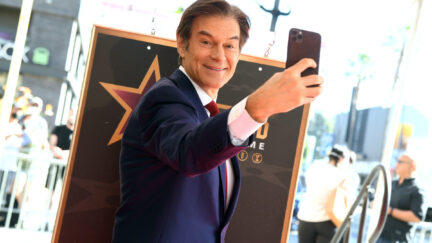 Dr Oz smiles at the camera as he holds his phone in front of himself to take a selfie.