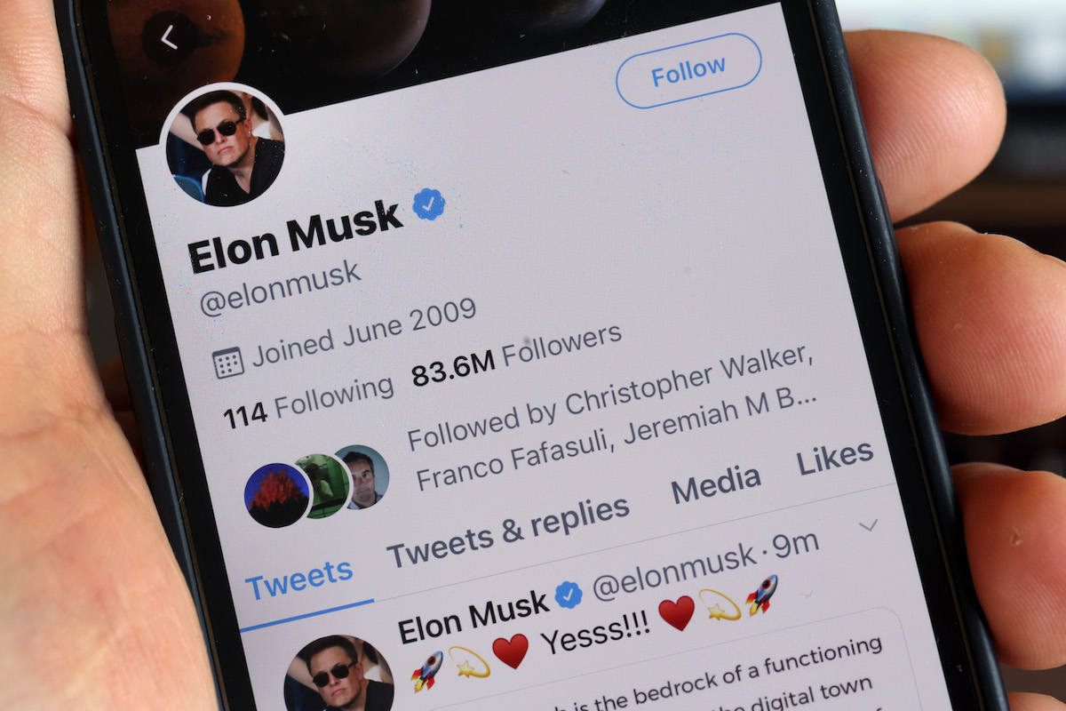 In closeup, a white hand holds a cell phone open to Elon Musk's Twitter profile.
