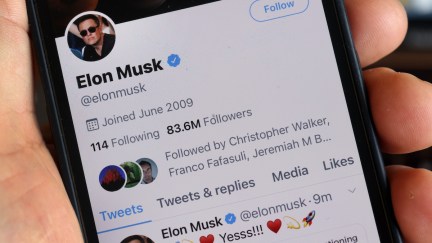 In closeup, a white hand holds a cell phone open to Elon Musk's Twitter profile.